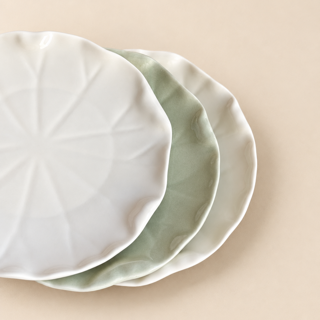 Three ceramic plates with scalloped edges stacked diagonally on a warm beige background. The top plate is white with a subtle radial starburst pattern, while the bottom two plates are a soft sage green.