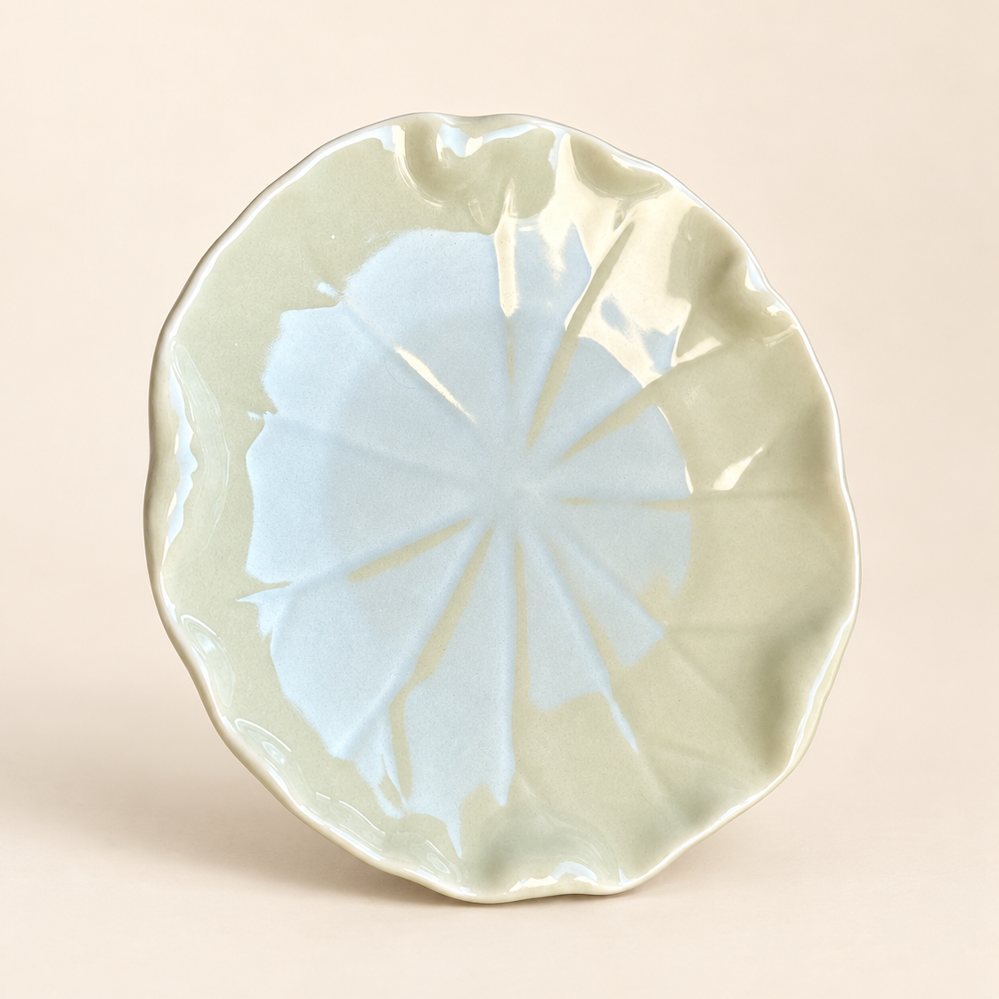 A ceramic shallow bowl with a scalloped, leaf-like rim and a glossy pale green glaze, featuring a light blue starburst pattern in the center, set against a solid beige background.
