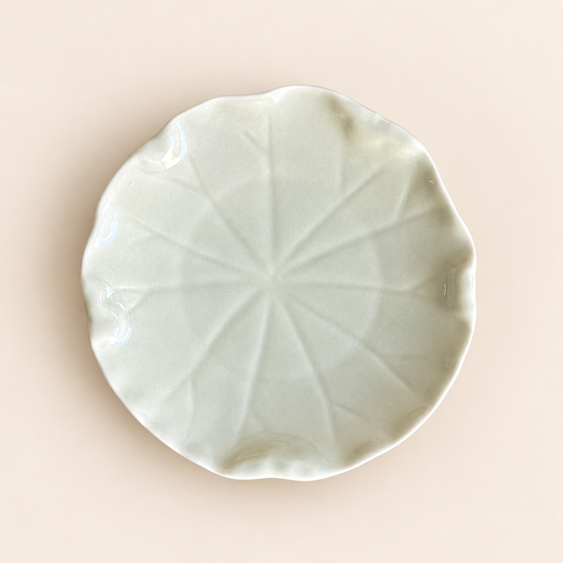 A small round ceramic dish with a scalloped edge and a leaf-like radial embossed pattern, shown from above on a soft beige background.