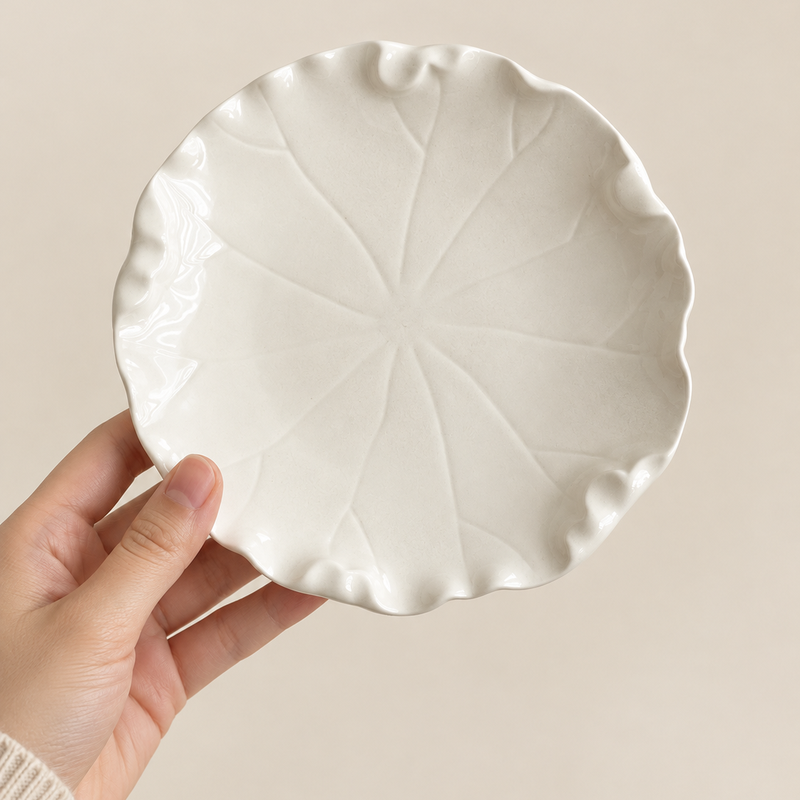 A hand holding a round white ceramic plate with a leaf-like scalloped edge and embossed radial leaf veins, shown against a plain beige background.