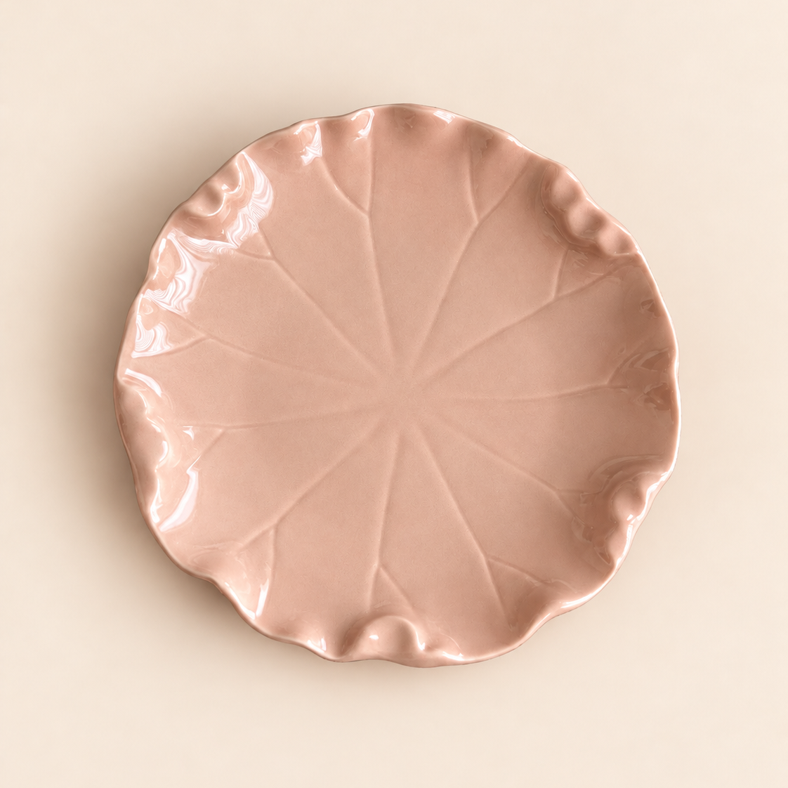 A round, leaf-shaped ceramic plate in a dusty rose pink color, featuring a glossy finish and a ruffled edge, photographed from directly above on a solid light beige background.
