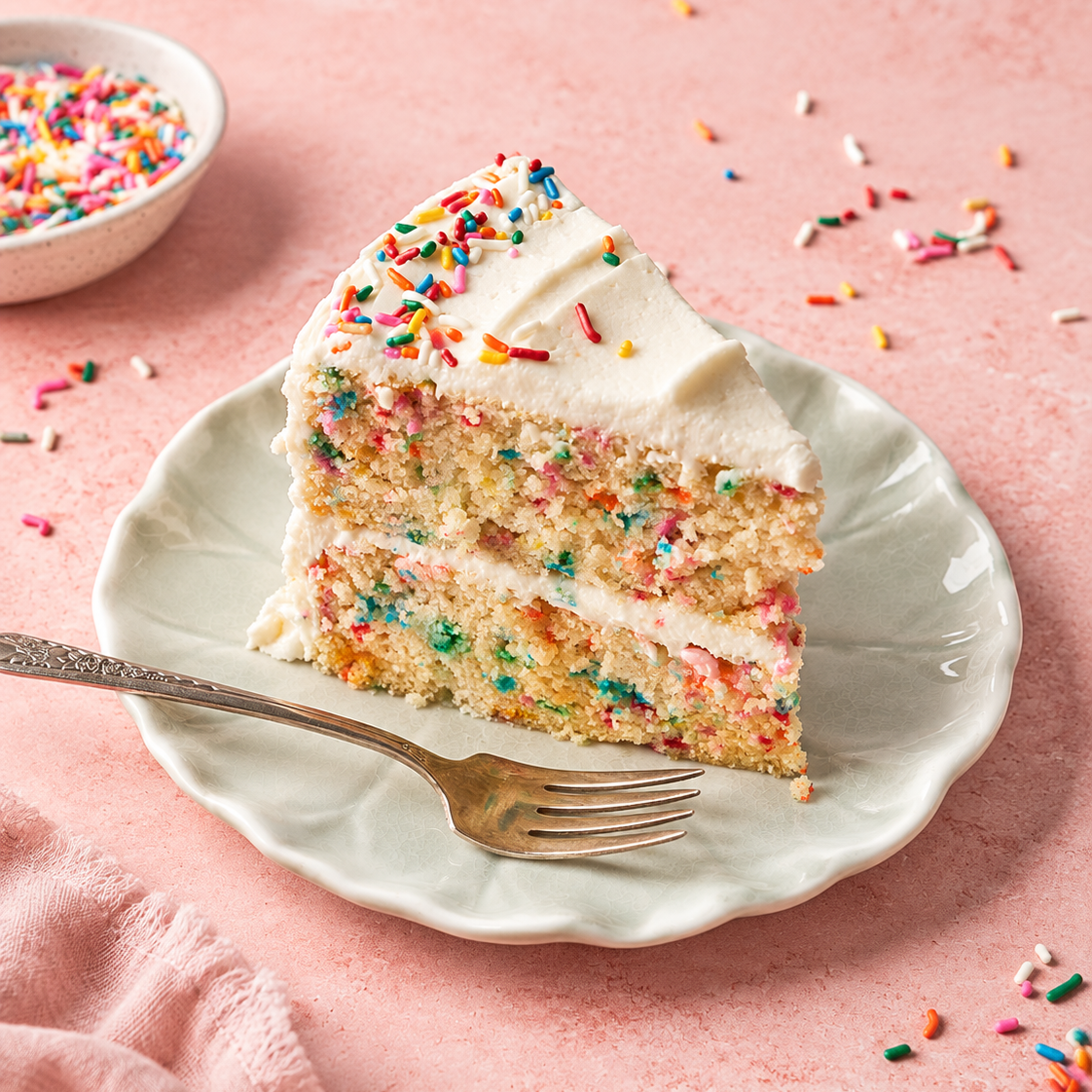 A slice of layered cake with white frosting and colorful sprinkles on a white plate, set on a pink surface with scattered sprinkles and a small bowl of sprinkles in the background.