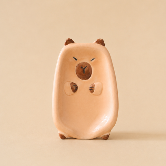A small ceramic figurine of a cute, rounded animal character soap dish in a warm peach-pink color with brown accents, standing on a solid beige background.