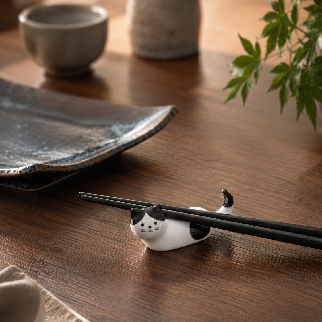 A small white cat-shaped chopstick rest with black markings, holding black chopsticks on a dark wooden table, with ceramic dishes and green leaves blurred in the background.