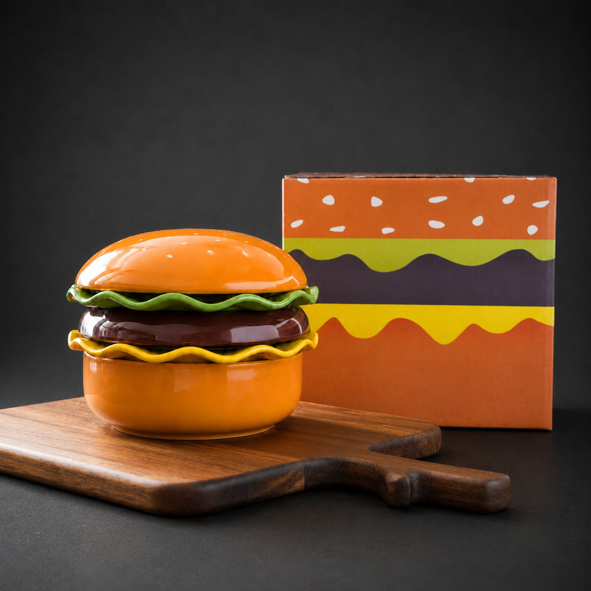 A ceramic hamburger-shaped container with an orange lid, green lettuce, brown patty, and yellow cheese details, displayed on a wooden cutting board with its illustrated orange box behind it against a dark background.