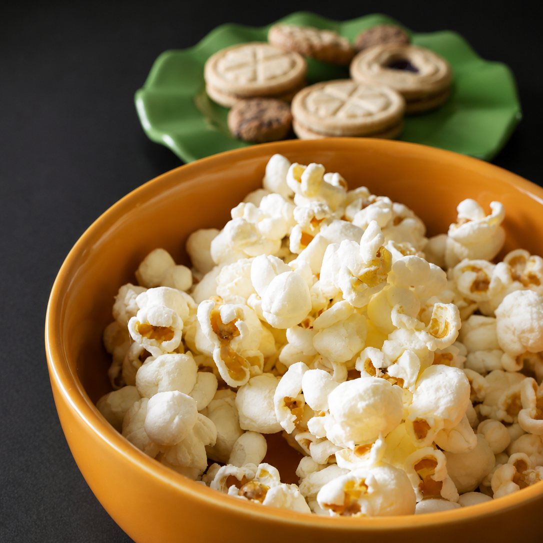 A yellow-orange ceramic bowl filled with fluffy white popcorn on a dark surface, with a green plate of cookies blurred in the background.