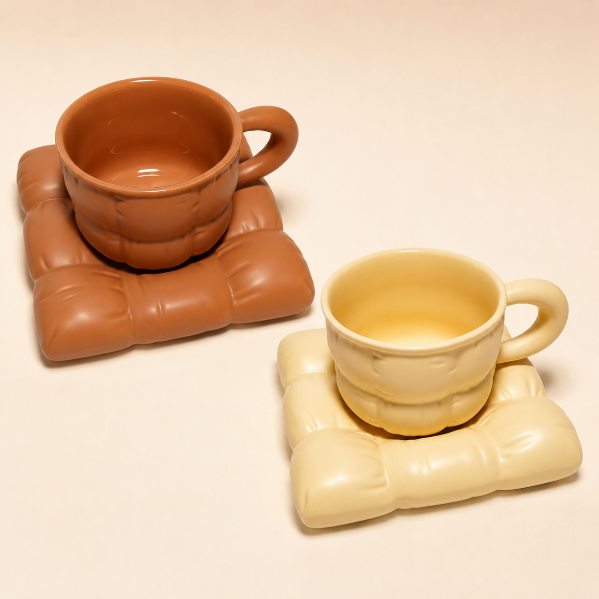 Two ceramic cups with matching sculptural coasters on a warm cream background. One cup and coaster are terracotta brown, and the other is pale cream yellow.