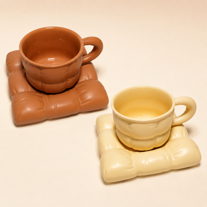 Two ceramic cups with matching sculptural coasters on a warm cream background. One cup and coaster are terracotta brown, and the other is pale cream yellow.
