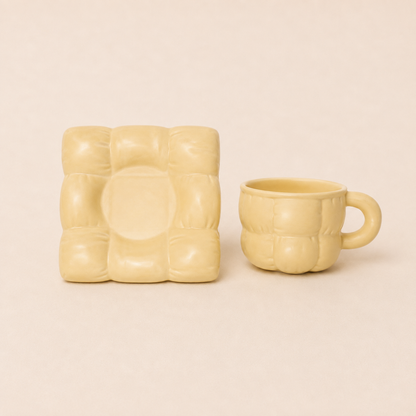 A pale yellow ceramic mug and matching square coaster with a raised grid pattern, set against a solid light beige background.