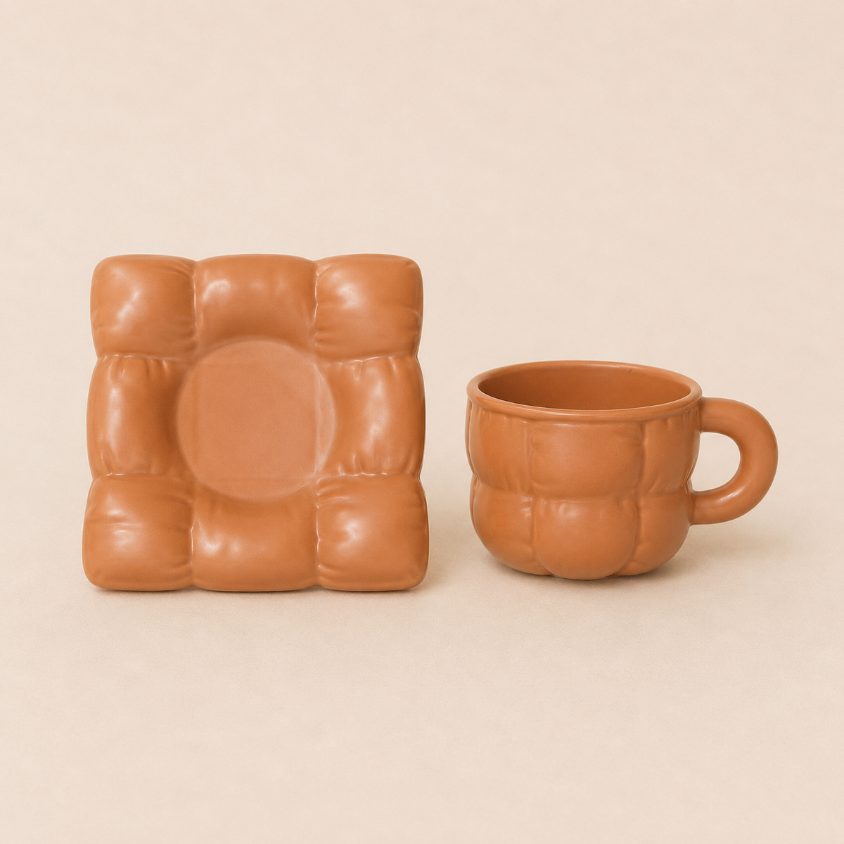 A terracotta-colored ceramic mug and matching square plate with a quilted, padded texture, displayed on a solid beige background.