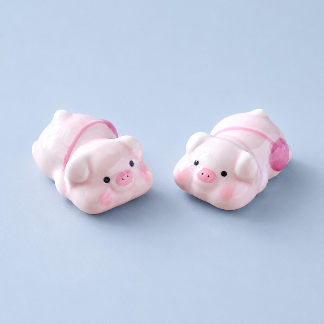 Two small pink pig-shaped ceramic decor or chopstick rests with black eyes and pink snouts, shown on a light blue background.