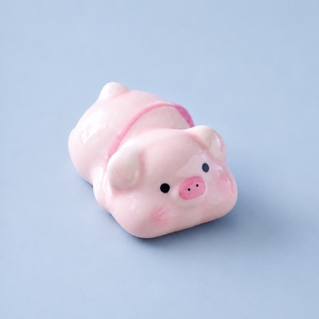 A small glossy pink pig figurine, chopstick rest or decorative object with black eyes and a pink snout, photographed on a smooth light blue background.