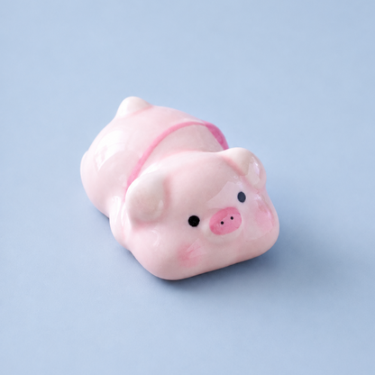 A small glossy pink pig figurine, chopstick rest or decorative object with black eyes and a pink snout, photographed on a smooth light blue background.