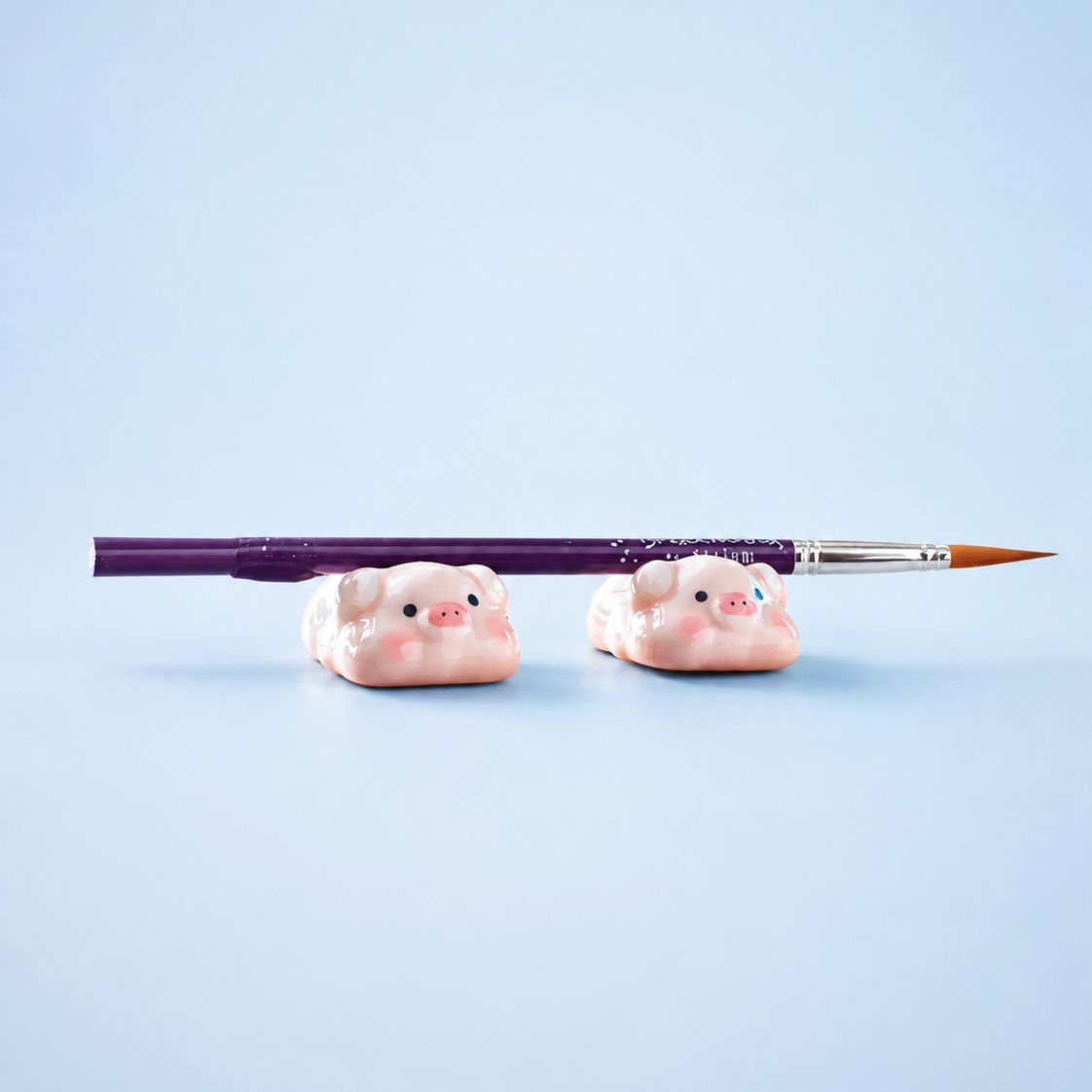 Two small pink pig-shaped desk or brushes holders on a light blue background, with a purple paintbrush resting horizontally across them.