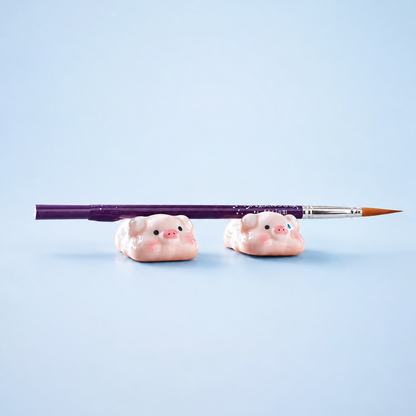 Two small pink pig-shaped desk or brushes holders on a light blue background, with a purple paintbrush resting horizontally across them.