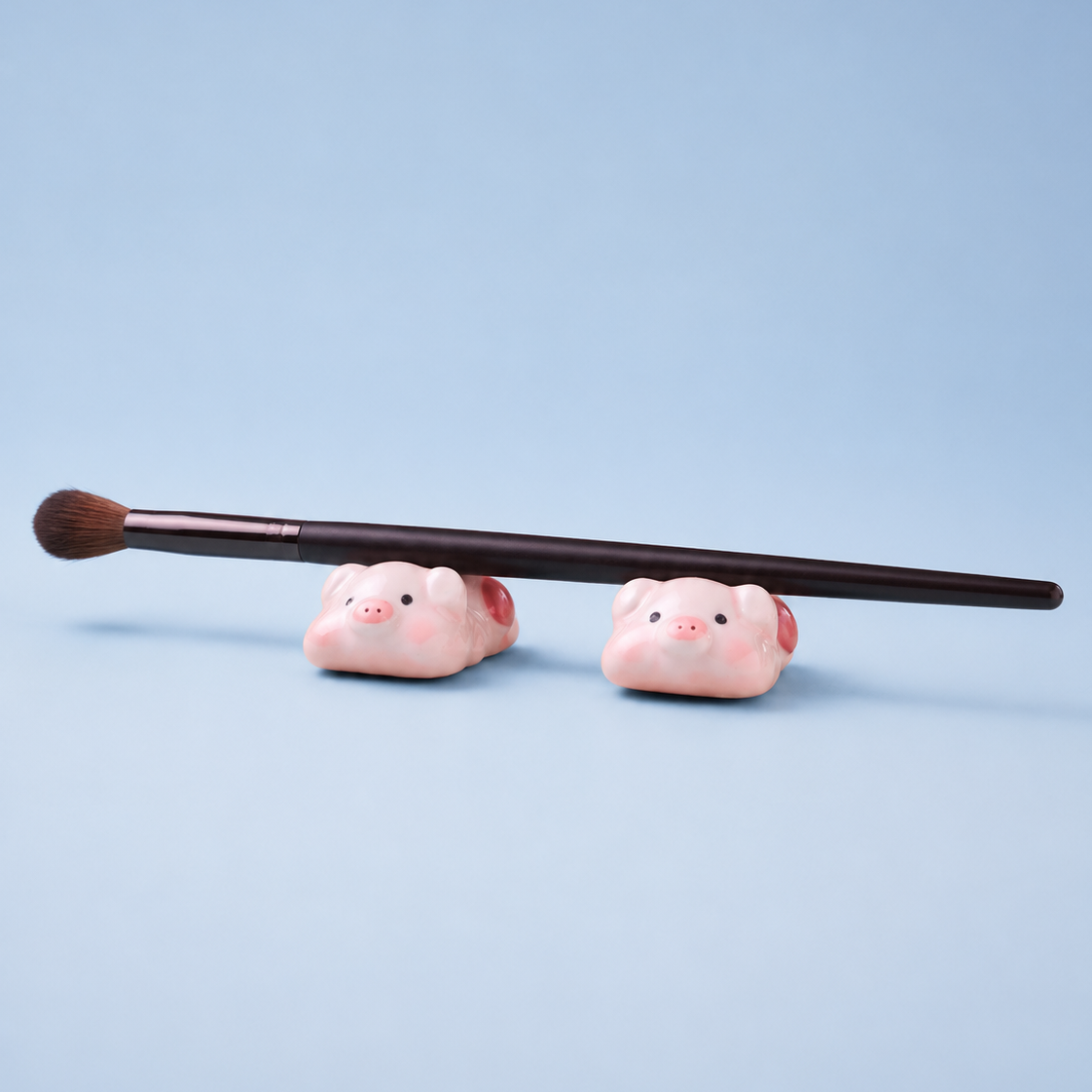 A makeup brush with a dark brown handle and brown bristles rests horizontally across two small pink pig figurines or chopstick rests on a light blue background.