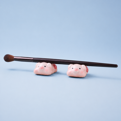A makeup brush with a dark brown handle and brown bristles rests horizontally across two small pink pig figurines or chopstick rests on a light blue background.