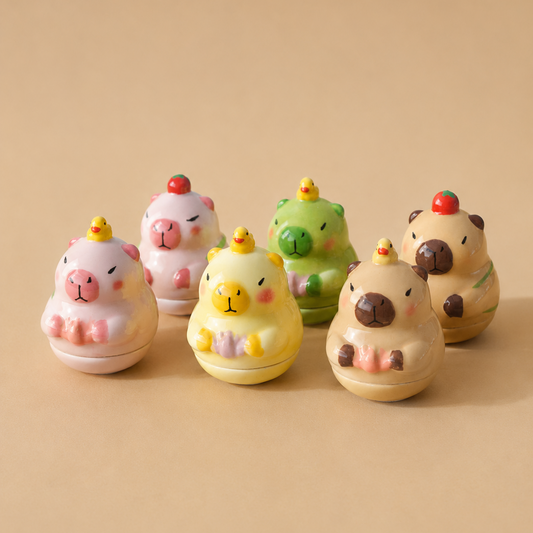 Six small capybara-shaped containers or toys in pastel colors, arranged in a row on a warm beige background. The capybaras are pink, white, green, and tan, each with a small yellow crown and a red cherry on top.