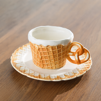 Biscuit-inspired ceramic mug with a waffle-textured body, white icing rim, pretzel-shaped handle, and matching ceramic saucer on a wooden table.