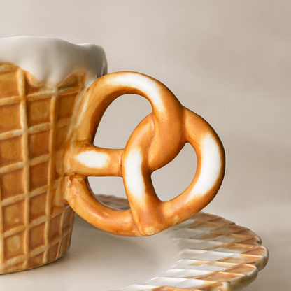 A close-up of a ceramic mug shaped like a pretzel, with a waffle-cone body, white pretzel-like handle, and white faux-foam top, shown on a warm beige background.