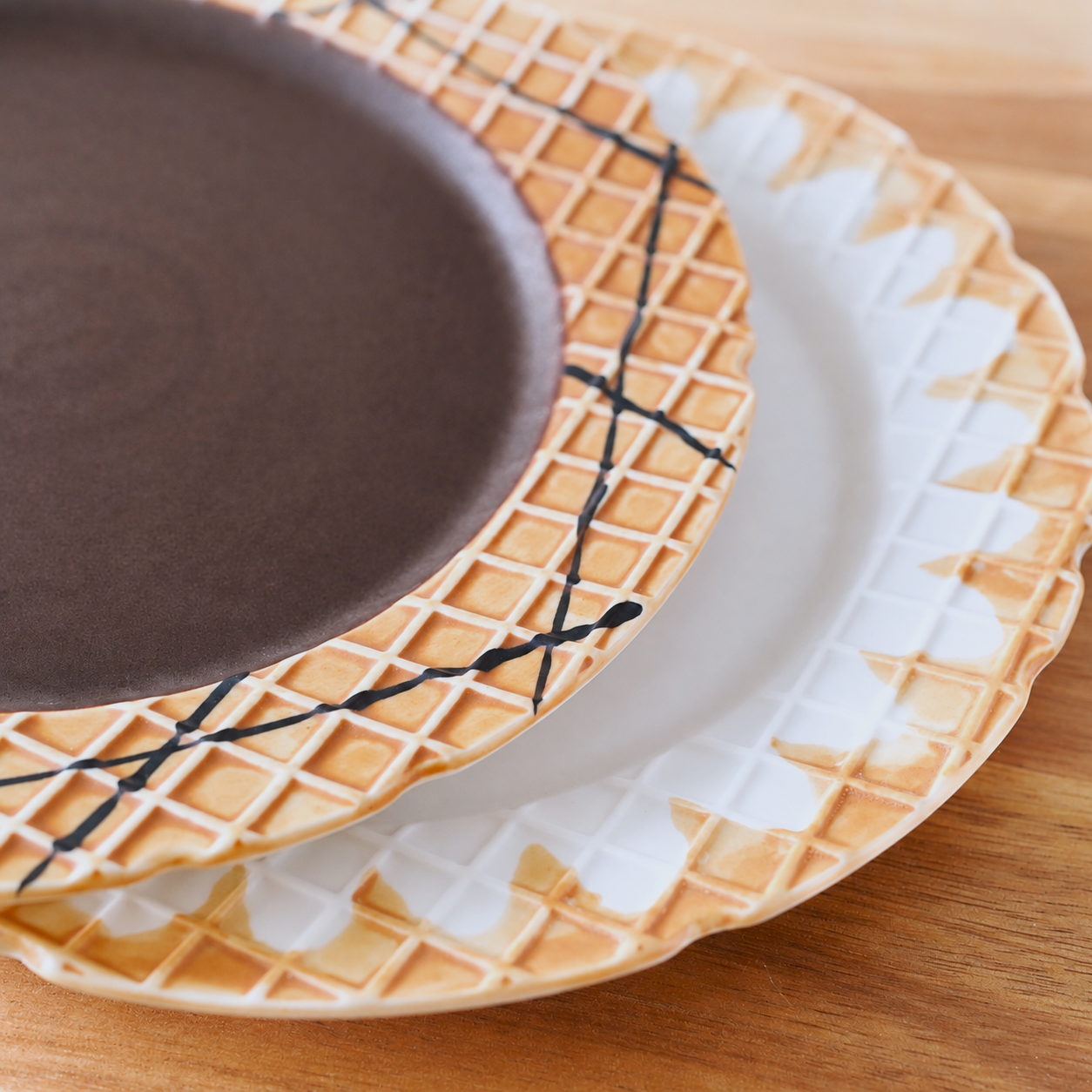 A close-up of a ceramic plate with a waffle pattern and a dark brown center, layered on top of another white plate with a waffle border, on a wooden surface.