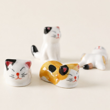 Four small ceramic cat figurines in different poses on a plain light beige background. The cats are painted white, yellow, black, and pink, with one lying down in the foreground and three standing or reclining behind it.