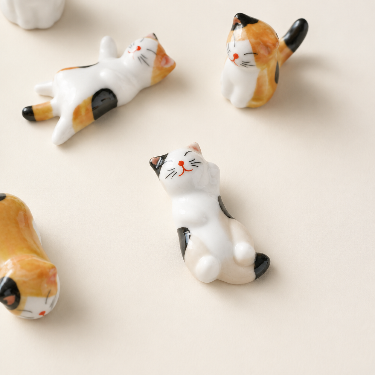 Several small ceramic cat figurines in calico colors are arranged on a plain warm off-white surface. The cats are posed casually, with one lying down and others sitting or standing, shown in a clean studio-style close-up.