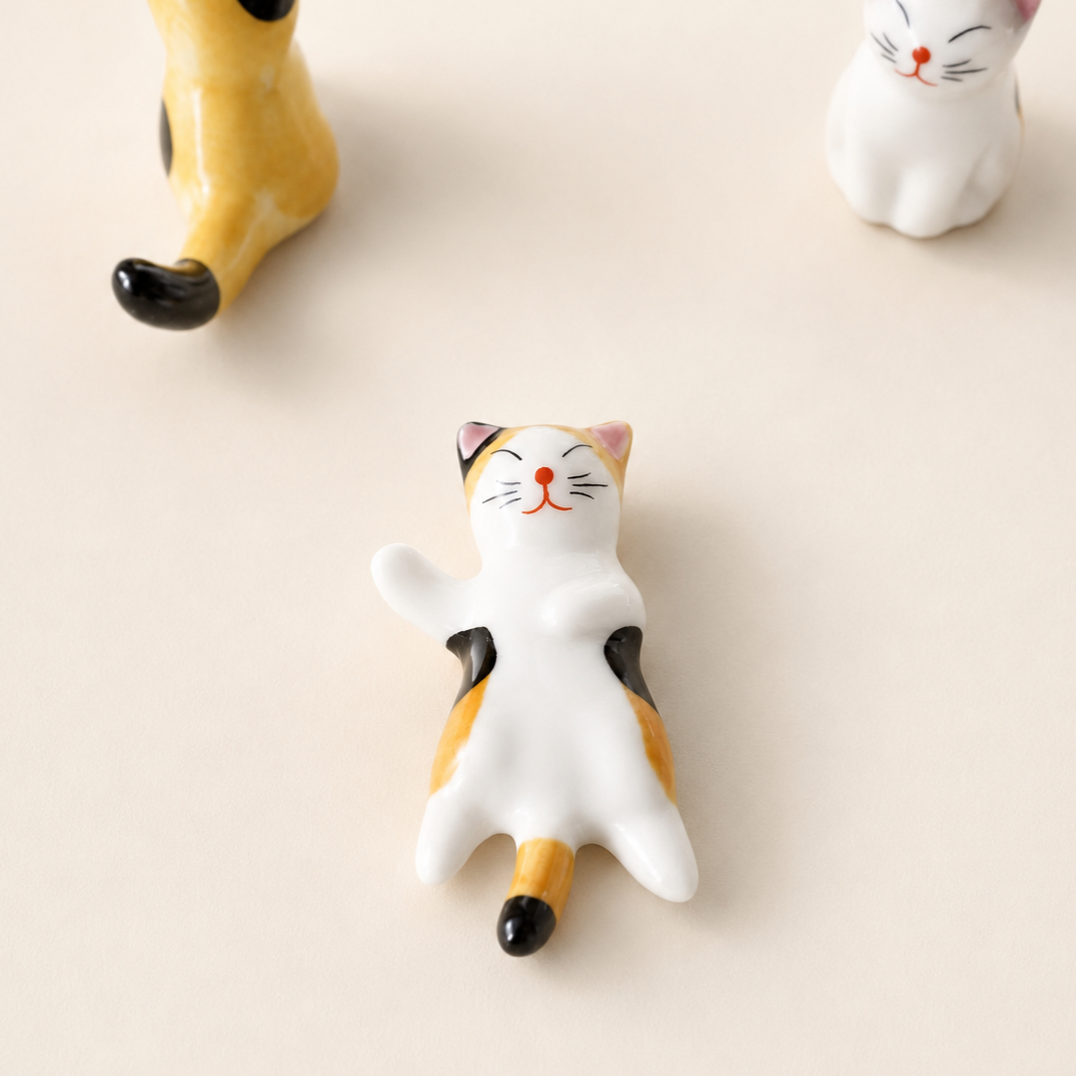 A small ceramic cat figurine lying on its back in a relaxed pose, with a white body, orange patches, black ears and tail tip, and a red nose, photographed on a warm off-white background. Part of another yellow cat figure appears at the top left, and another white cat figure appears at the top right.