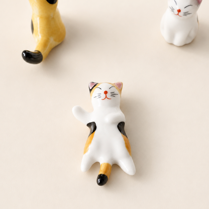 A small ceramic cat figurine lying on its back in a relaxed pose, with a white body, orange patches, black ears and tail tip, and a red nose, photographed on a warm off-white background. Part of another yellow cat figure appears at the top left, and another white cat figure appears at the top right.
