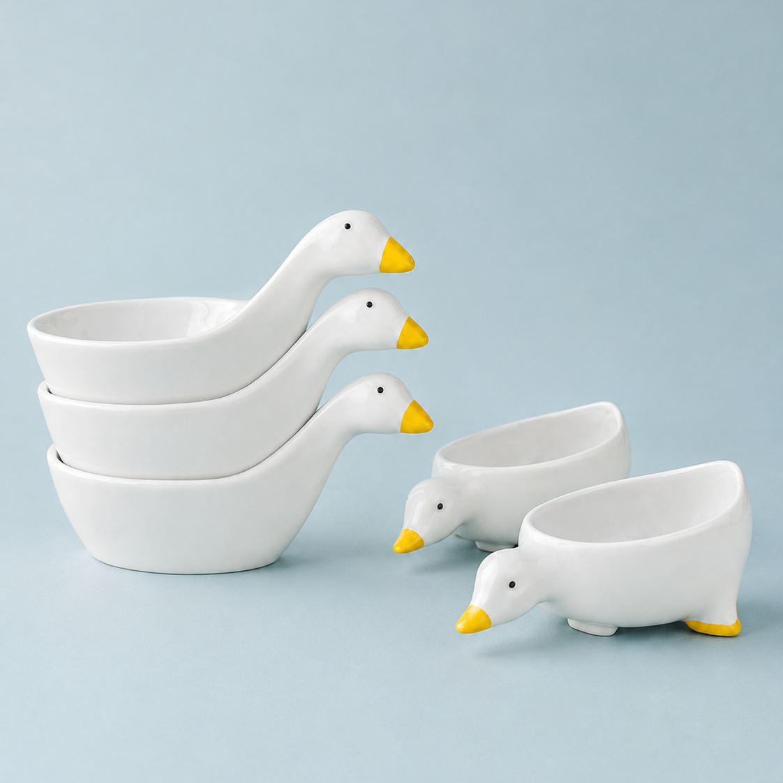 A set of ceramic duck-shaped bowls in white with yellow beaks and feet, arranged on a light blue background.