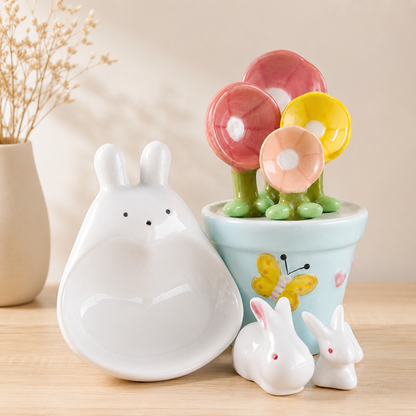 A ceramic rabbit-shaped dish in white sits beside a small light blue flower pot with colorful flower-shaped lids in pink, yellow, and peach, plus two small white rabbit figurines on a wooden surface.