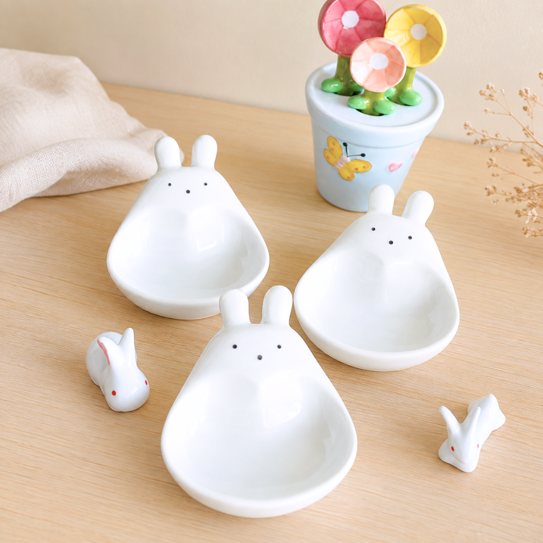 Three white ceramic bunny-shaped bowls arranged on a light wood tabletop, with two small matching bunny figurines beside them and a small floral pot in the background.