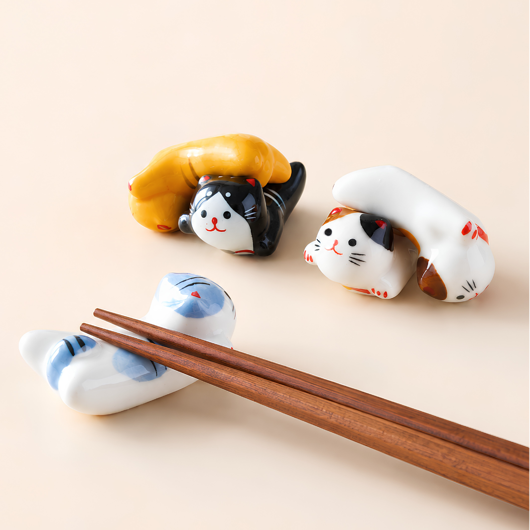 Three ceramic cat-shaped chopstick rests in white, black, and calico colors, with a pair of wooden chopsticks resting on one of the cat holders against a warm beige background.