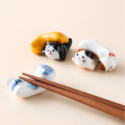 Three ceramic cat-shaped chopstick rests in white, black, and calico colors, with a pair of wooden chopsticks resting on one of the cat holders against a warm beige background.