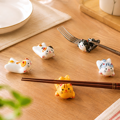 Five small ceramic cat-shaped chopstick holders in assorted colors arranged on a wooden table, with a fork resting on one holder and chopsticks resting on another.