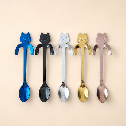 Five metallic spoons shaped like cats, shown in blue, black, silver, gold, and rose gold finishes against a solid beige background.