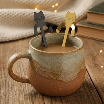 Two cat-shaped metal tea stirrers, one black and one gold, resting in a ceramic mug filled with iced coffee on a wooden table.