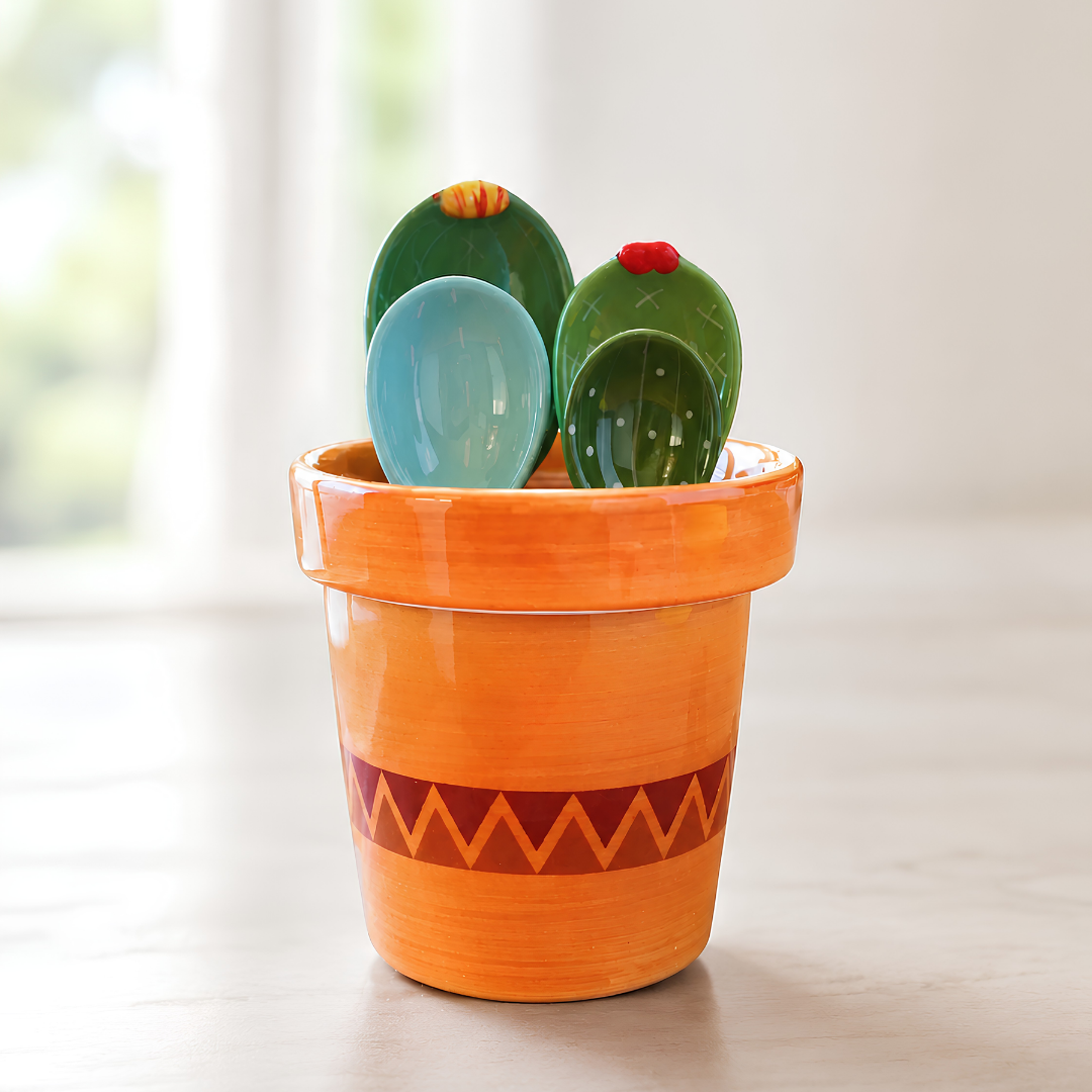A ceramic measuring spoon in bright orange with a red zigzag pattern, holding three measuring spoons shaped like cacti in green, blue, and light blue, set on a light wooden surface with a soft-focus background.