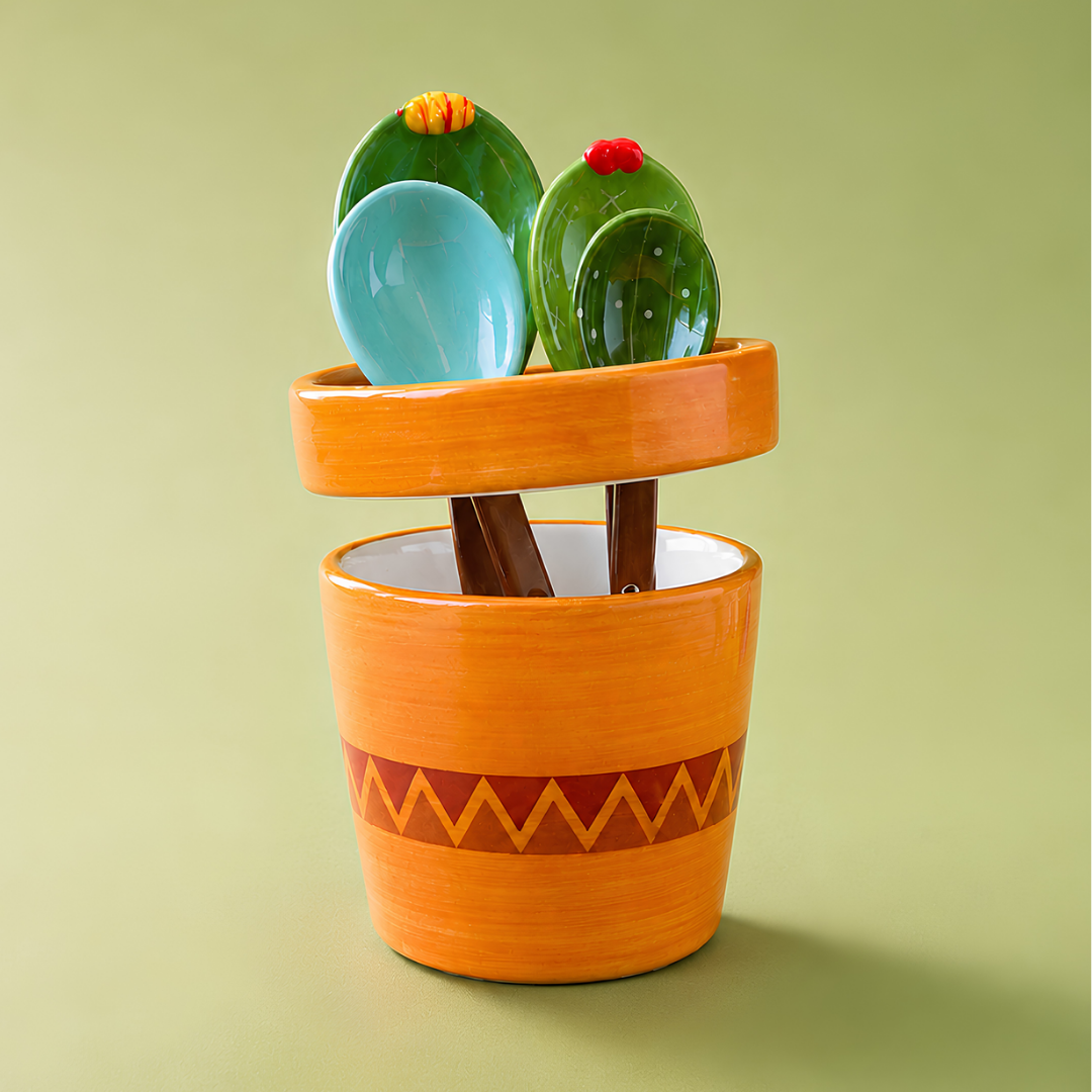 A ceramic cactus-themed utensil holder, measuraing cup  and spoon set on a solid light green background. The orange pot-shaped holder features a zigzag pattern and contains green cactus-shaped spoons and a light blue spoon.