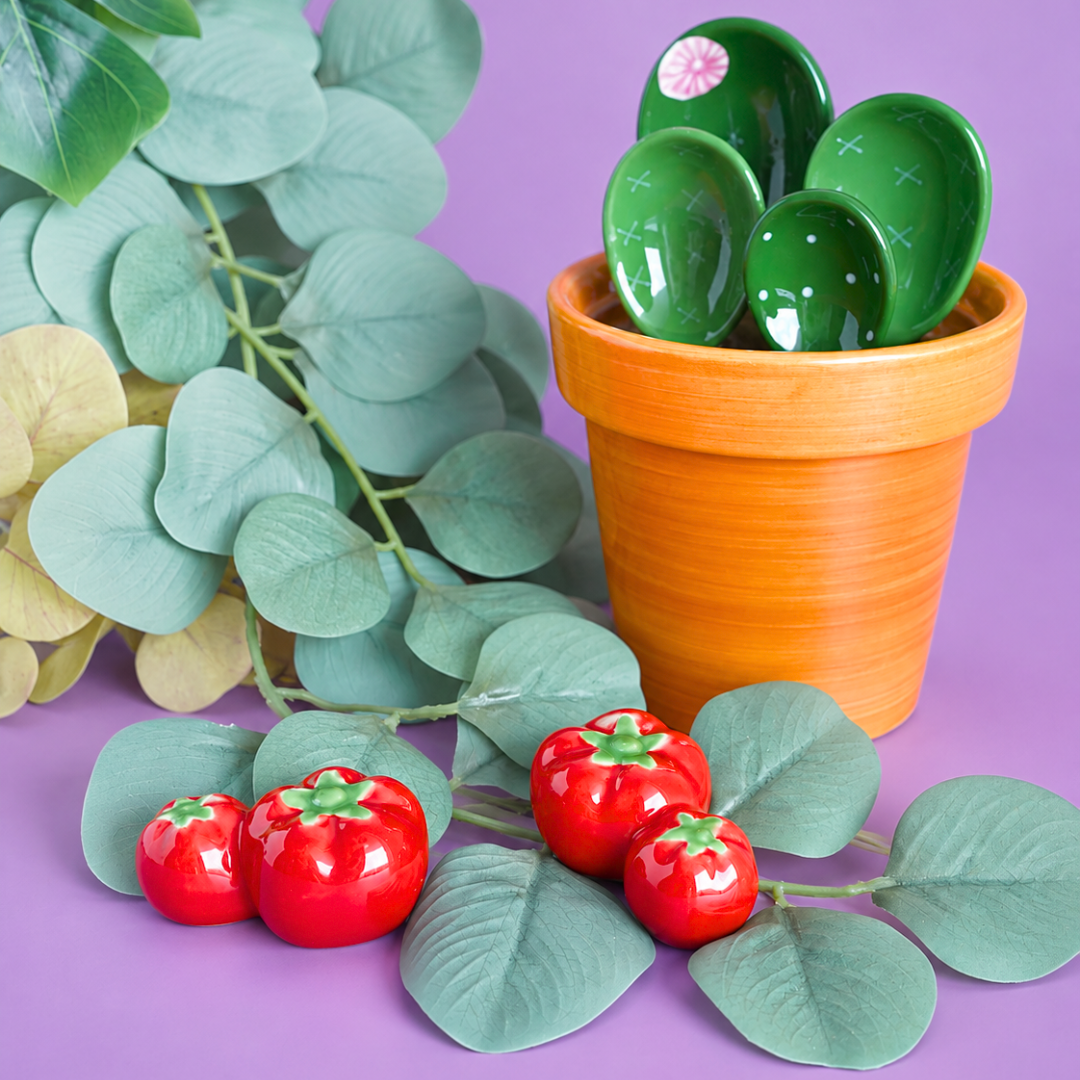 A decorative ceramic cactus planter holding four glossy green cactus-shaped plates, styled with faux eucalyptus leaves and red tomato ceramic decor or chopstick rest on a solid purple background.