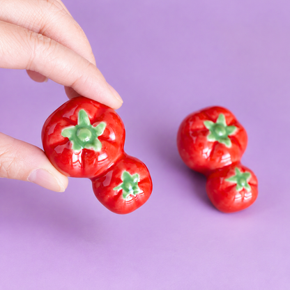 A hand holds a pair of glossy red tomato-shaped ceramic decor or chopstick rest with green stem details against a solid lavender background. Two matching earrings are displayed on the right.