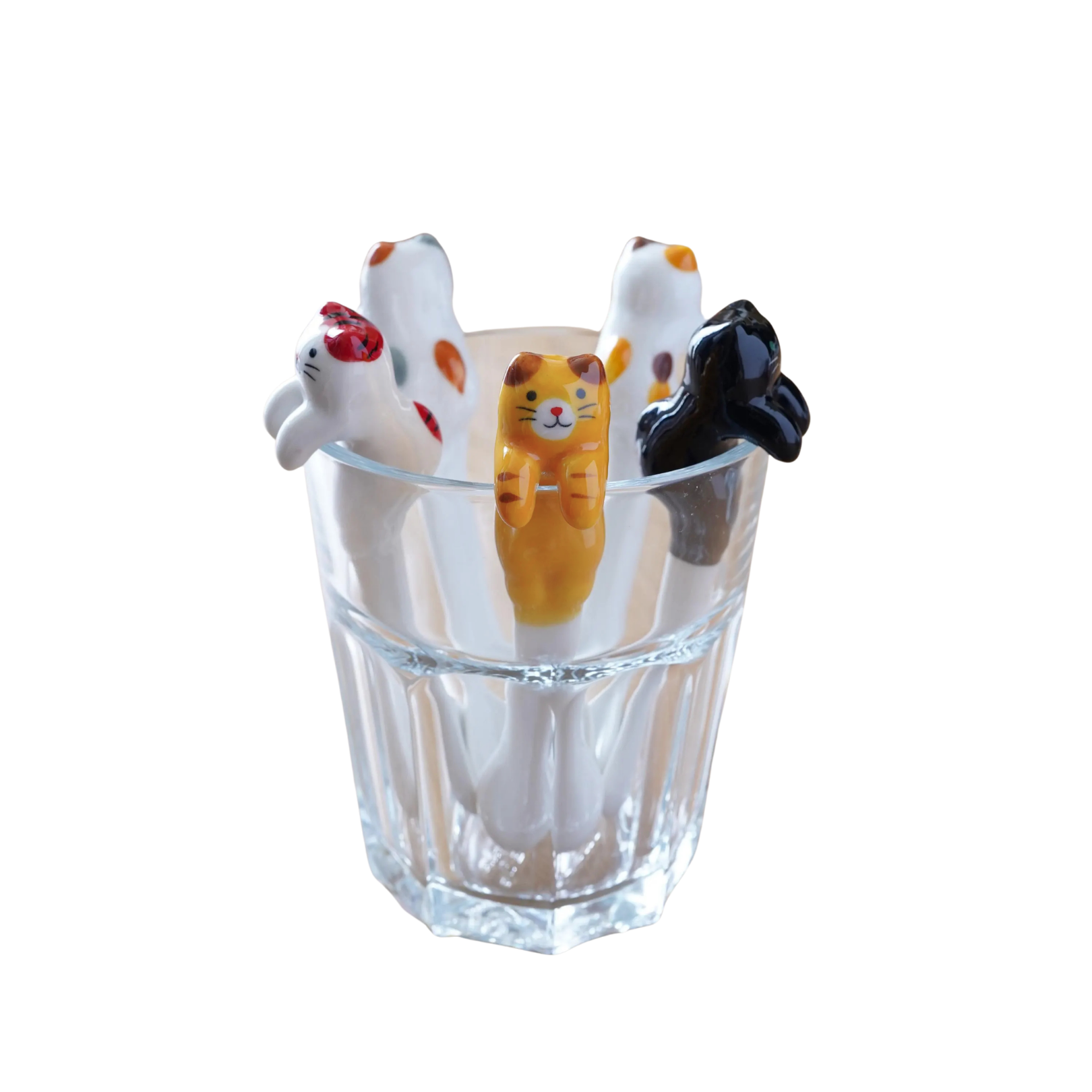A clear ribbed glass cup holding four small cat-shaped figurines in white, orange, and black, photographed on a plain white background.