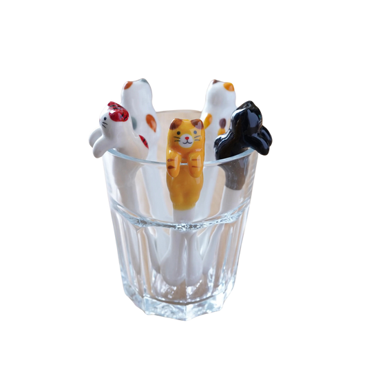 A clear ribbed glass cup holding four small cat-shaped figurines in white, orange, and black, photographed on a plain white background.
