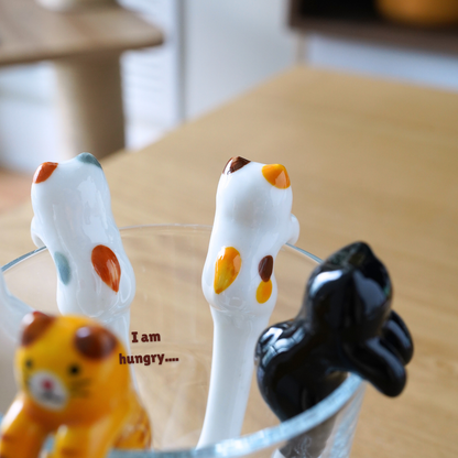 Three ceramic cat-shaped spoons in white, black, and yellow sitting in a clear glass bowl on a wooden table, with the text 'I am hungry.