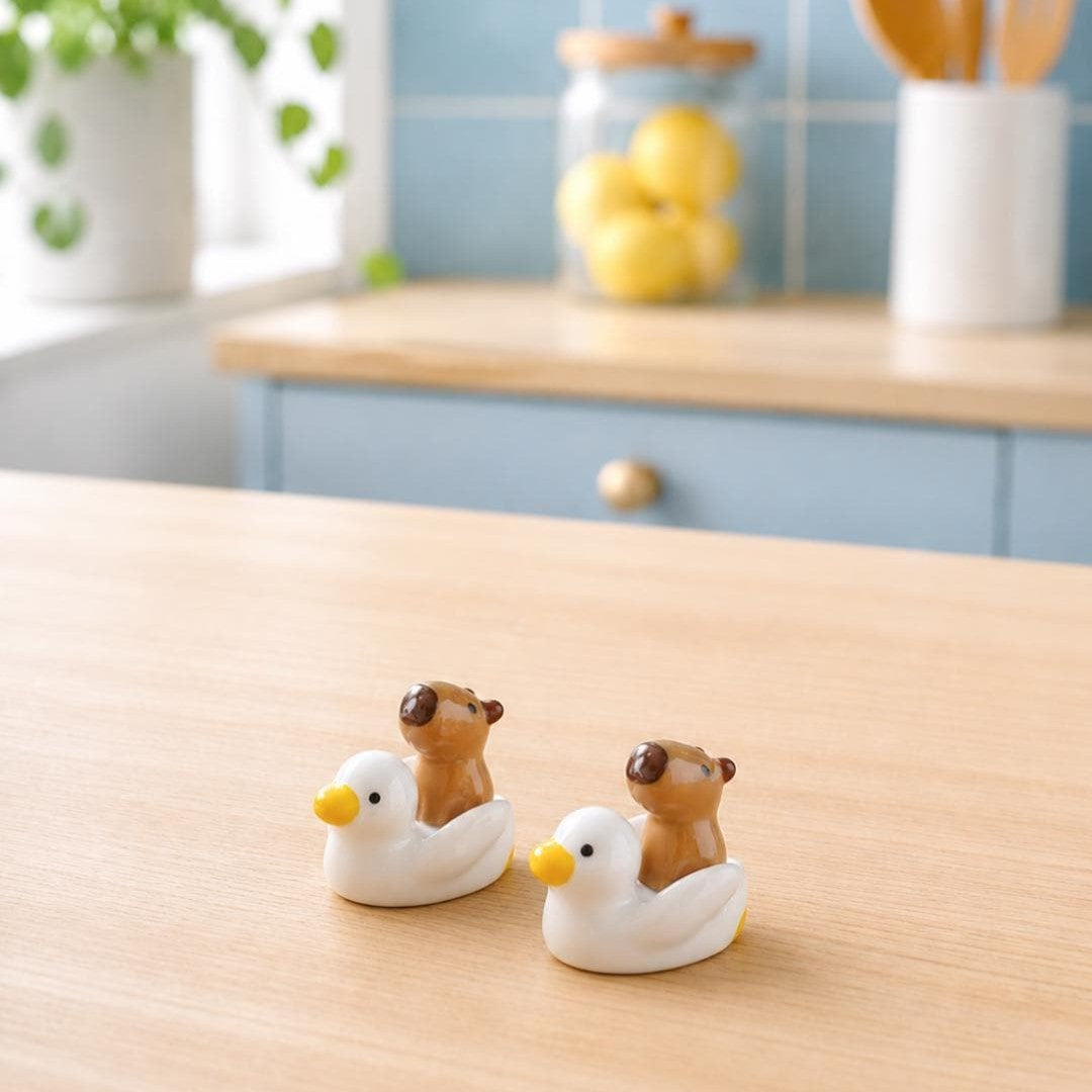 Two small ceramic duck figurines with brown bear riders, placed on a light wood tabletop in a softly blurred kitchen setting.